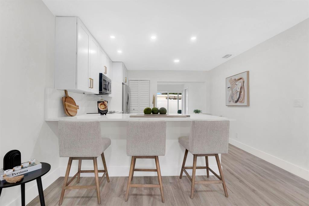 A modern white kitchen features a breakfast bar with three stools and light wood flooring — 1115 Stillwater Drive, Jupiter FL 33458, JUPITER, FL