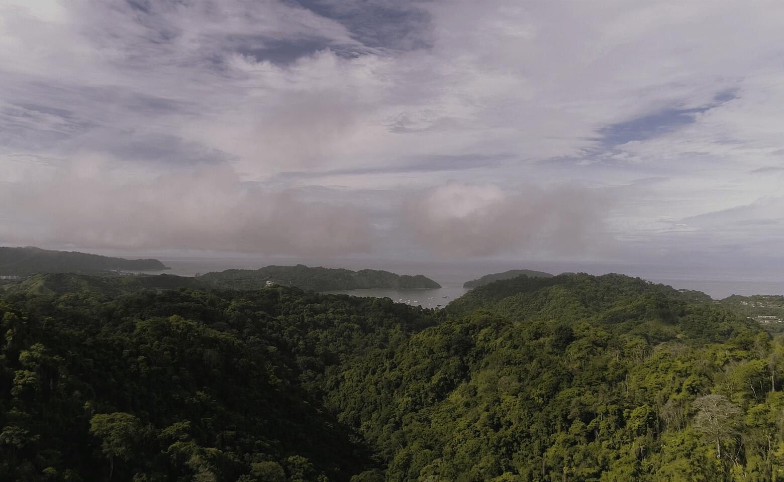 An aerial view shows a dense green forest overlooking a bay with boats at sea — 0 Playa Caletas, Canton De Garabito, Puntarenas, FL