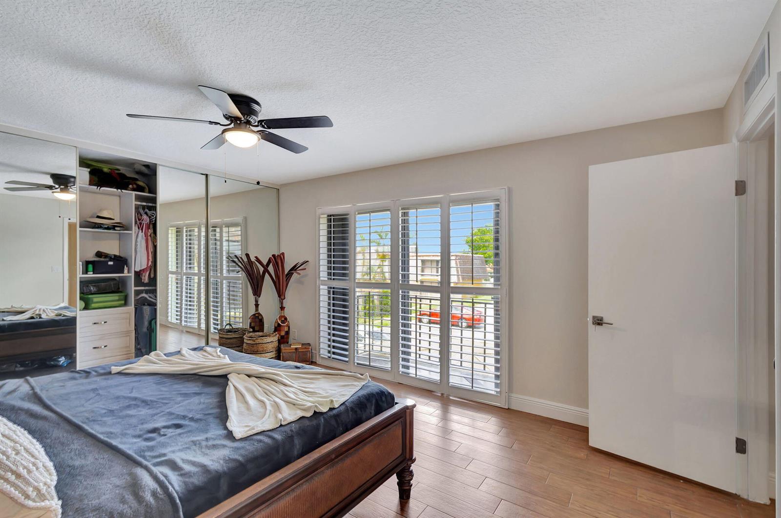 A bedroom features wood-look tile flooring, a mirrored closet, a ceiling fan, and plantation shutters — 99 Lake Arbor Drive, Palm Springs FL 33461, PALM SPRINGS, FL