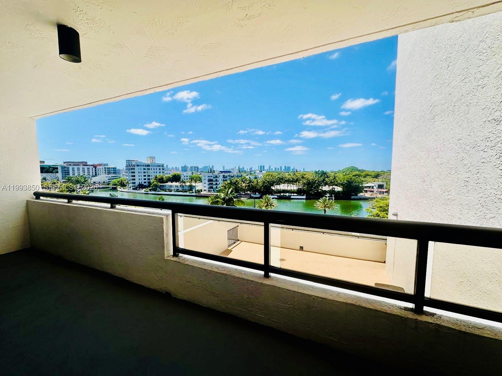A balcony overlooks a canal, lush trees, and a distant city skyline under a blue sky — 2555 Collins Ave # 704, Miami Beach FL 33140, MIAMI BEACH, FL