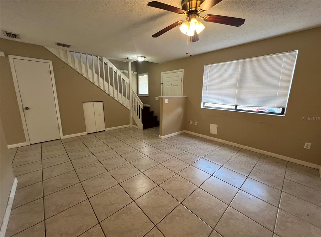 A living room features beige tile flooring, a staircase with white railings, and a ceiling fan — 100 E BAYWOOD SQUARE, DAYTONA BEACH, FL