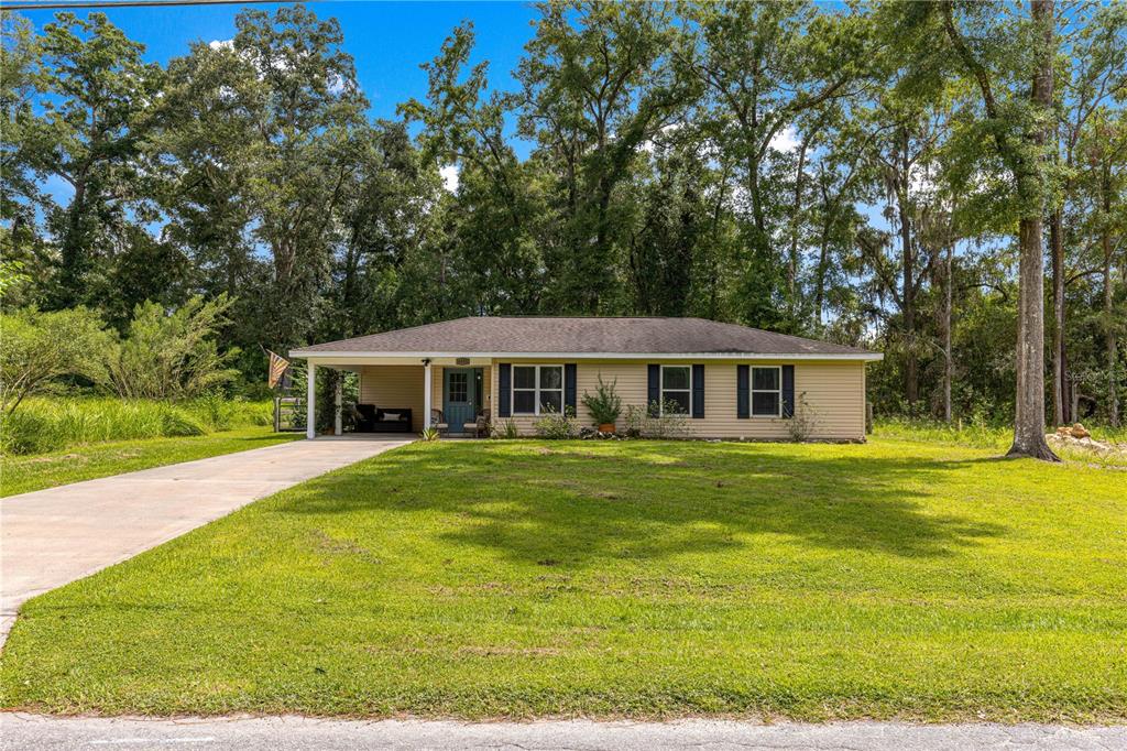 A single-story house with tan siding, black shutters, and a carport sits on a grassy lot — 6353 NW 52ND AVENUE, OCALA, FL