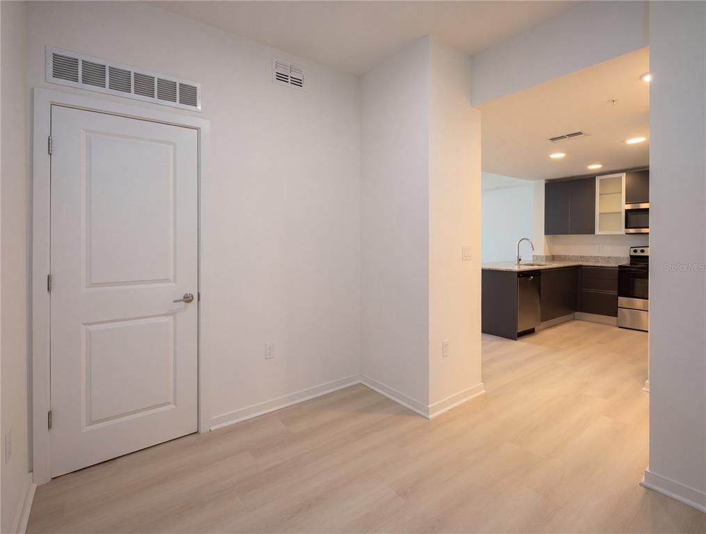 An interior view shows a white-walled room with light wood flooring leading to a kitchen — 5579 MILLENIA PARK BOULEVARD UNIT 217, ORLANDO, FL