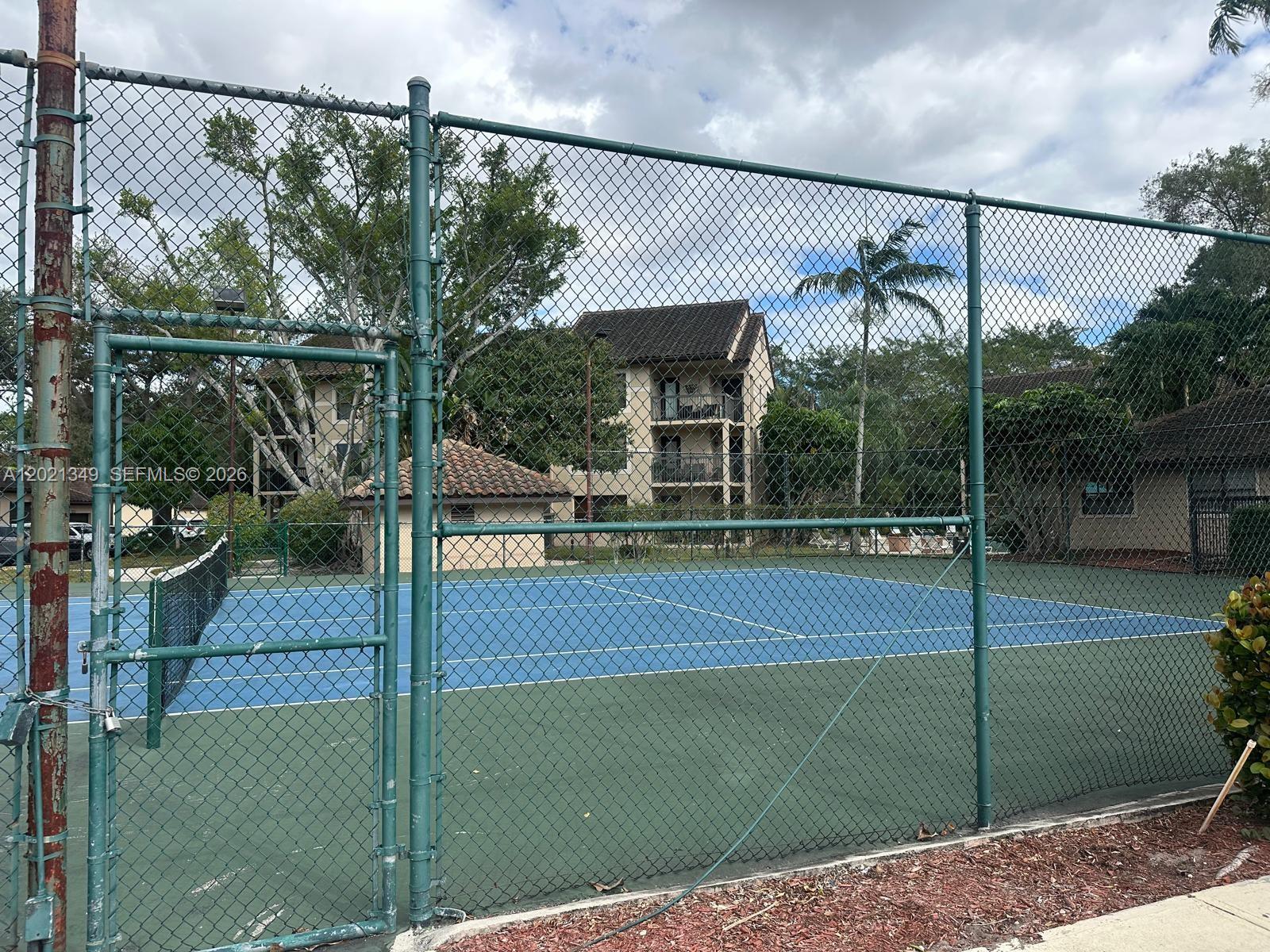 A blue tennis court is enclosed by a chain-link fence with apartment buildings in the background — 4481 W Mcnab Rd # 37, Pompano Beach FL 33069, POMPANO BEACH, FL