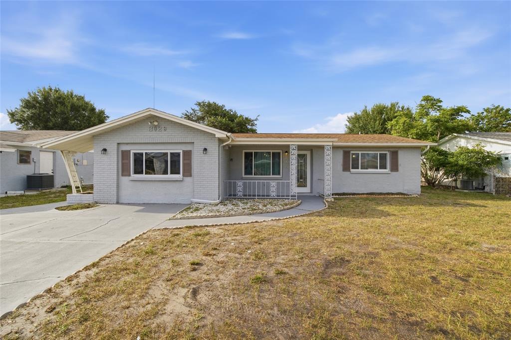 A single-story gray brick house features a carport, a front porch, and a grassy yard — 3029 HUNTINGTON ROAD, HOLIDAY, FL