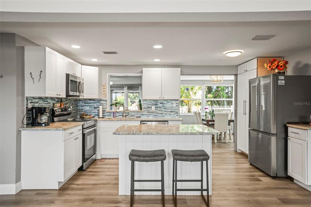 A kitchen features white cabinets, granite countertops, a central island with bar stools, and stainless appliances — 4704 KOALA DRIVE, HOLIDAY, FL