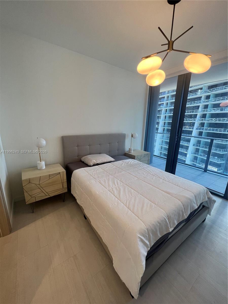 A bedroom features a gray tufted headboard, geometric nightstands, and a modern chandelier near a balcony — 1000 Brickell Plaza # 2701, Miami FL 33131, MIAMI, FL