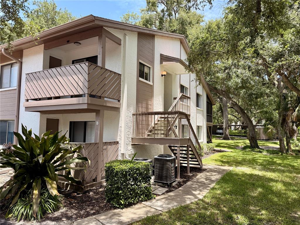 A two-story apartment building exterior features a wooden balcony, outdoor staircase, and surrounding trees — 2363 FLANDERS WAY UNIT D, SAFETY HARBOR, FL