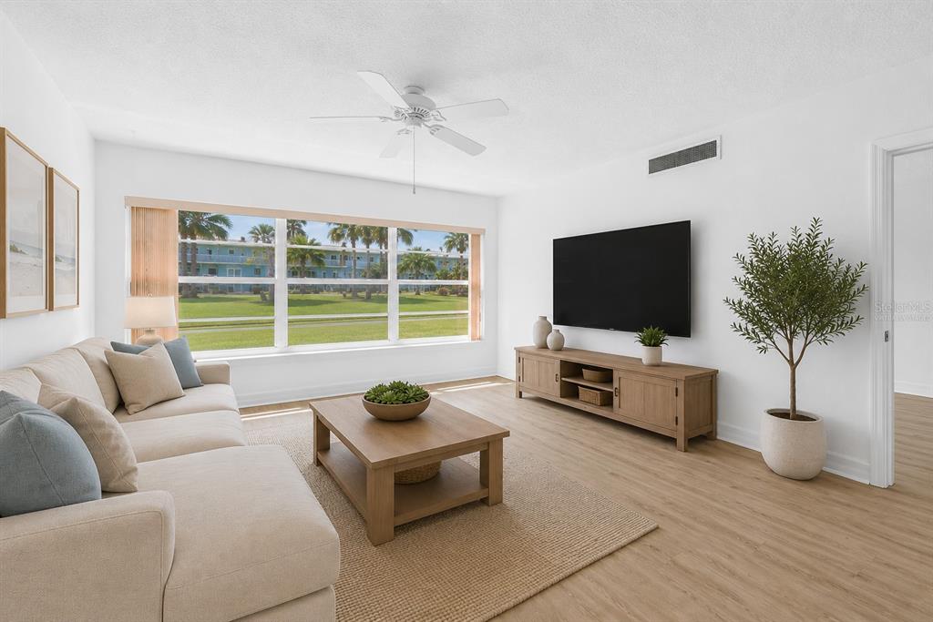 A bright living room features a beige sofa, wooden coffee table, media console, and large windows — 300 64TH AVENUE UNIT 117, ST PETE BEACH, FL