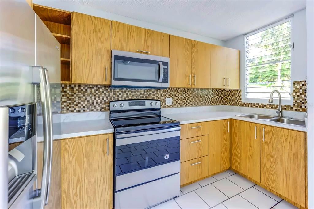 A kitchen features light wood cabinets, stainless steel appliances, a mosaic backsplash, and white countertops — Alturas de Piedras Blanca UNITS D-202, GUAYNABO, PR