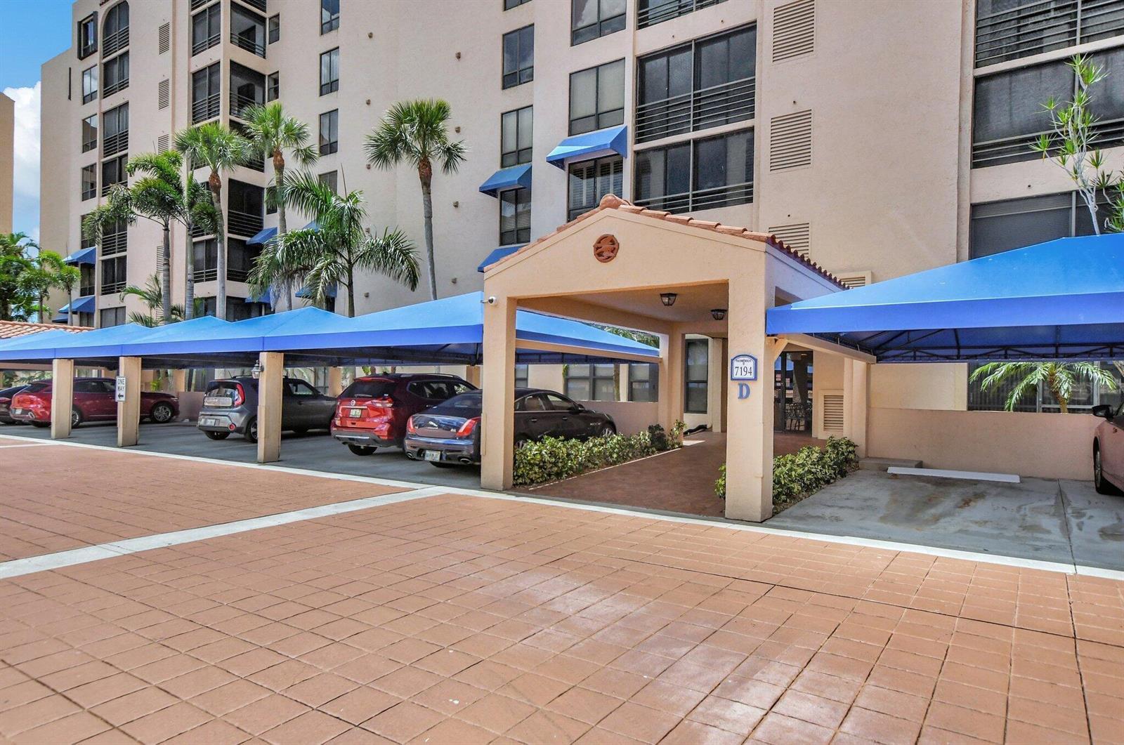 A covered entrance with a blue awning leads to a multi-story building with parking spaces — 7194 Promenade Drive # 102, Boca Raton FL 33433, BOCA RATON, FL