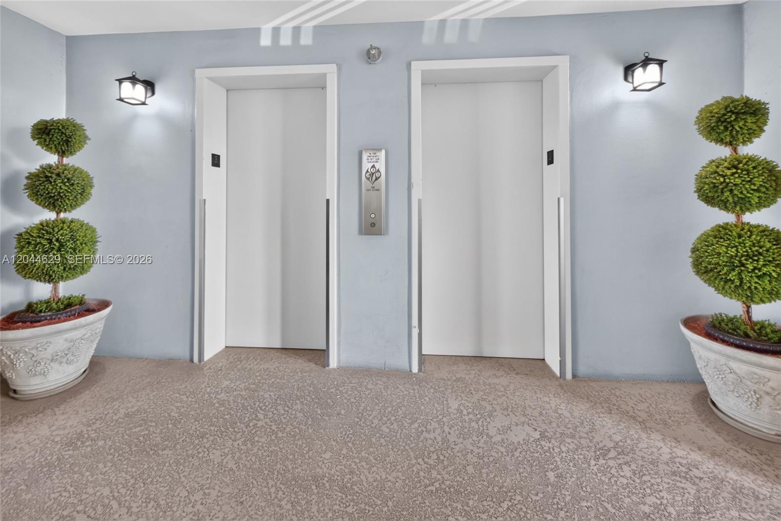 Two white elevator doors are centered between two potted topiary trees on a textured floor — 9101 W Sample Rd # 306, Coral Springs FL 33065, CORAL SPRINGS, FL