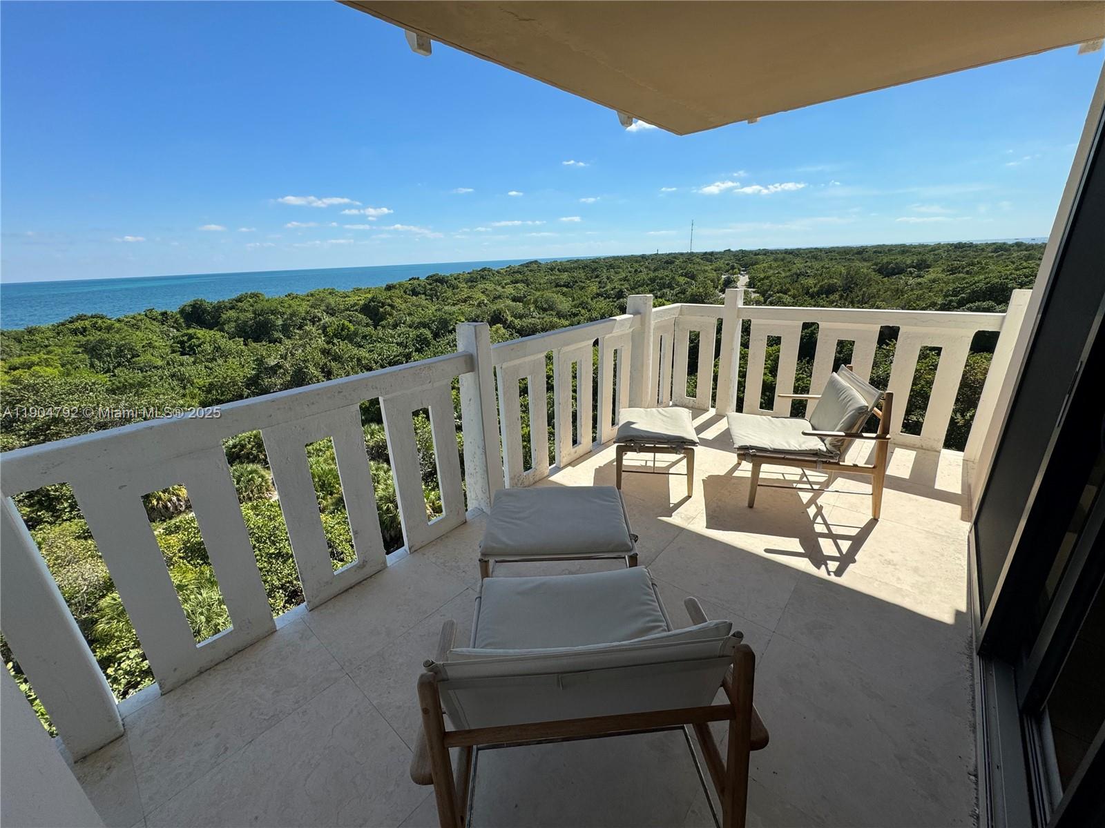 A balcony with two chairs and ottomans overlooks a dense forest and the ocean — 1121 Crandon Blvd # D608, Key Biscayne FL 33149, KEY BISCAYNE, FL