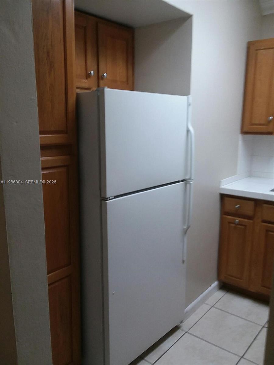 A white top-freezer refrigerator is positioned between wooden kitchen cabinets and a tiled floor — 140 Royal Palm Rd # 107, Hialeah Gardens FL 33016, HIALEAH GARDENS, FL