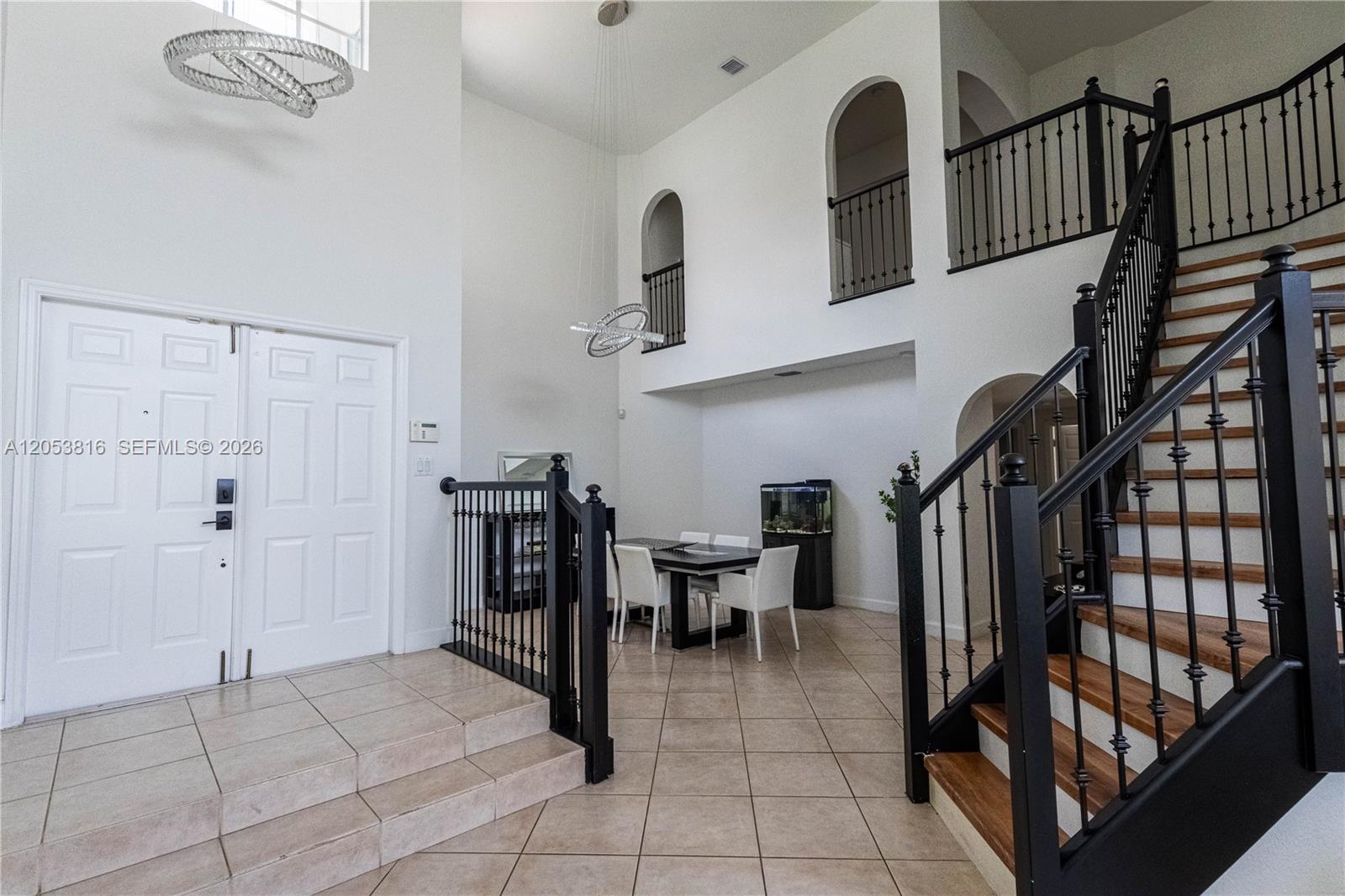 A high-ceilinged foyer features a dining set, tiled floors, and a wooden staircase — 10873 NW 71st St, Doral FL 33178, DORAL, FL