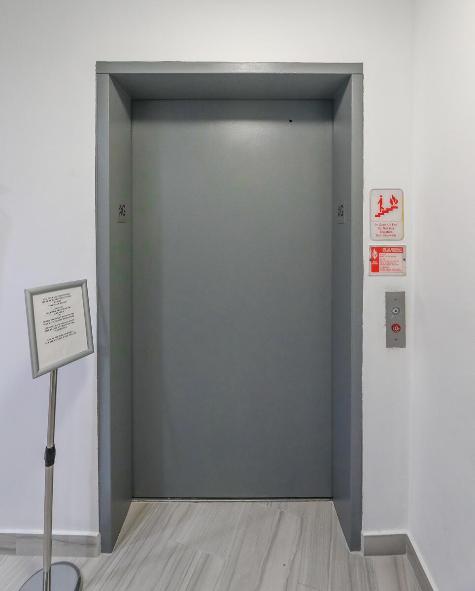A gray elevator door is set into a white wall with signage and call buttons — 2100 Brickell Avenue # 406, Miami FL 33129, MIAMI, FL