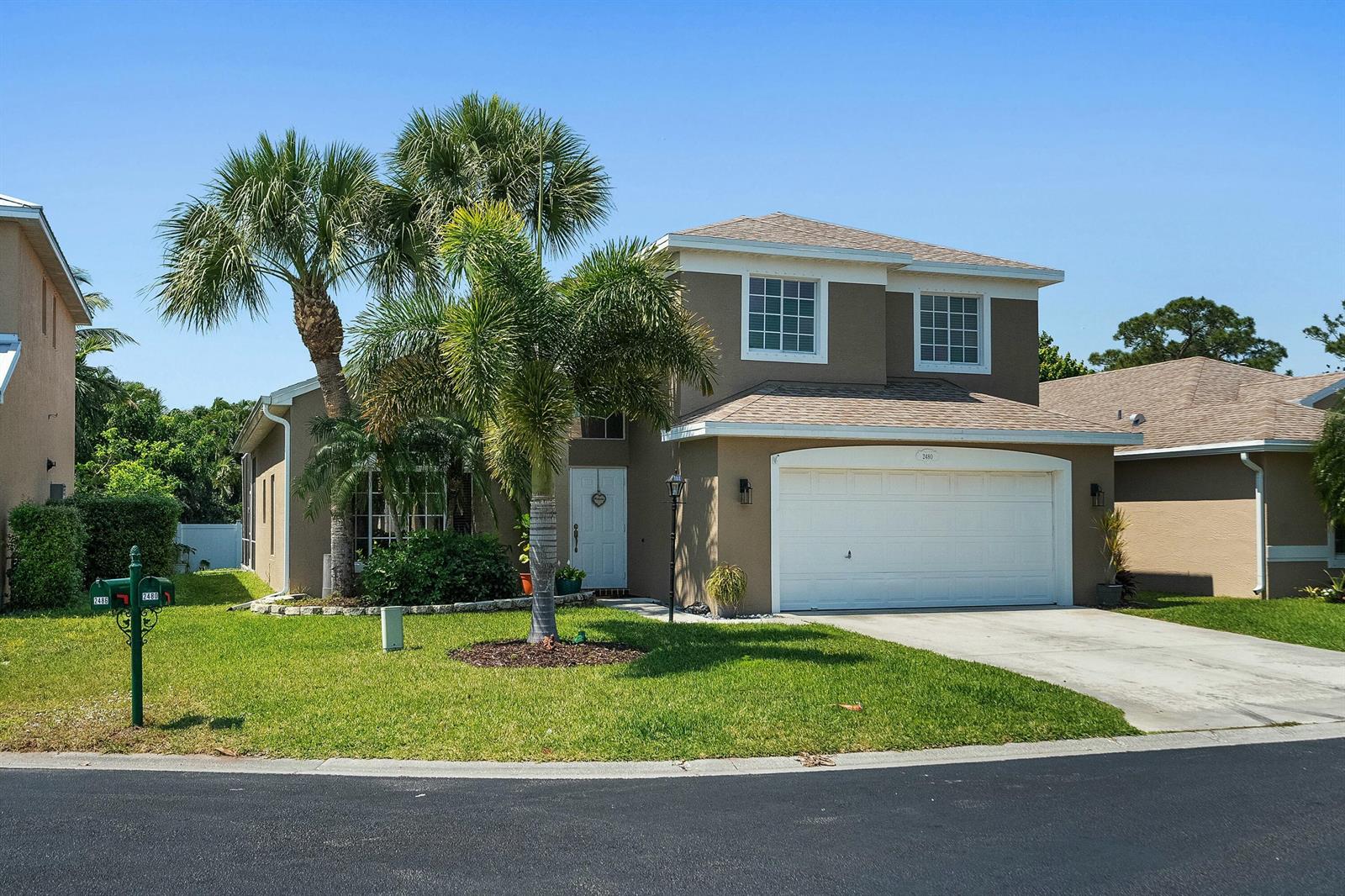 A two-story tan house with a white garage door and palm trees in the front yard — 2480 SE Springtree Place, Stuart FL 34997, STUART, FL