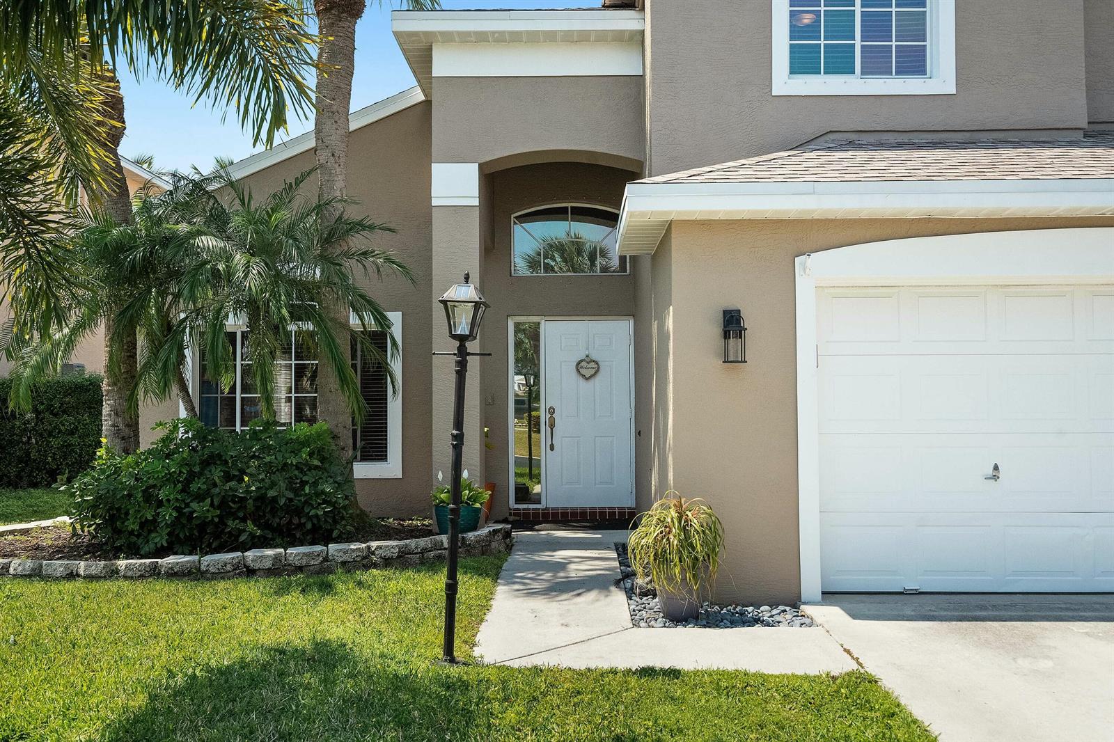 A tan two-story house features a white front door, a garage, and a palm tree — 2480 SE Springtree Place, Stuart FL 34997, STUART, FL