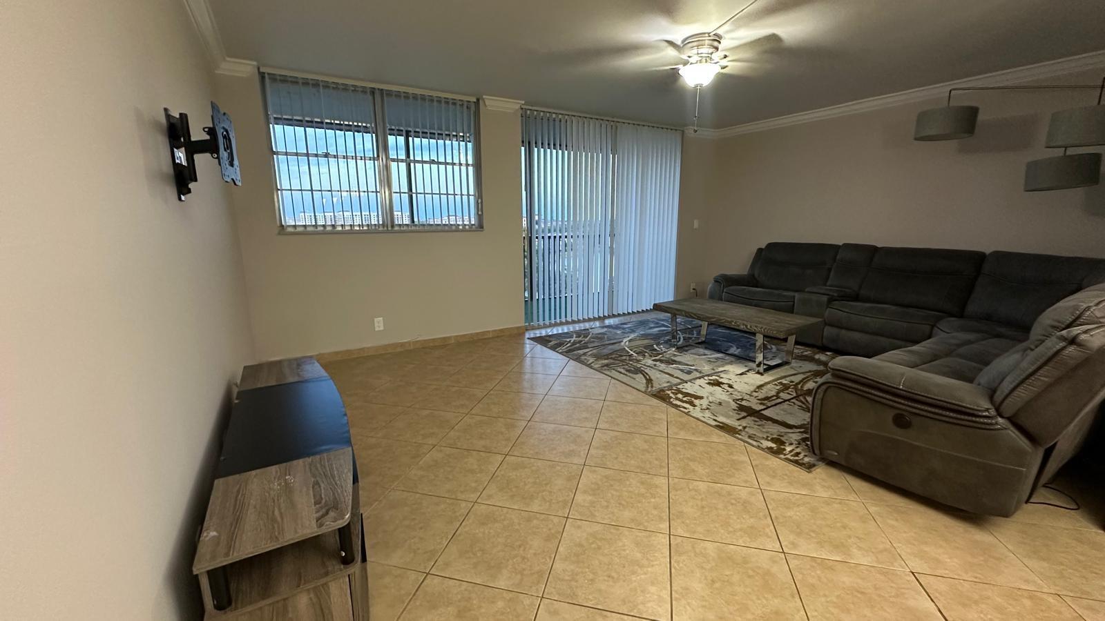 A living room features tile flooring, a gray sectional sofa, and a wall-mounted television bracket — 3300 W Rolling Hills Circle # 708, Davie FL 33328, DAVIE, FL
