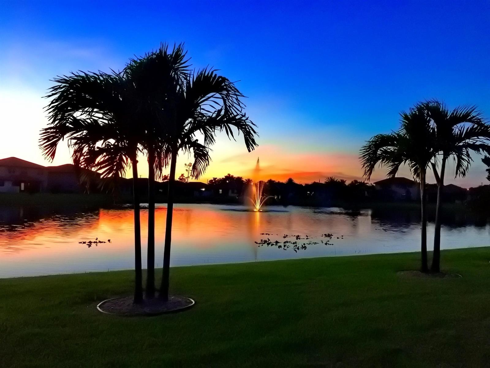 A sunset view over a lake with a fountain, palm trees, and surrounding residential homes — 7171 Prudencia Drive, Lake Worth FL 33463, LAKE WORTH, FL