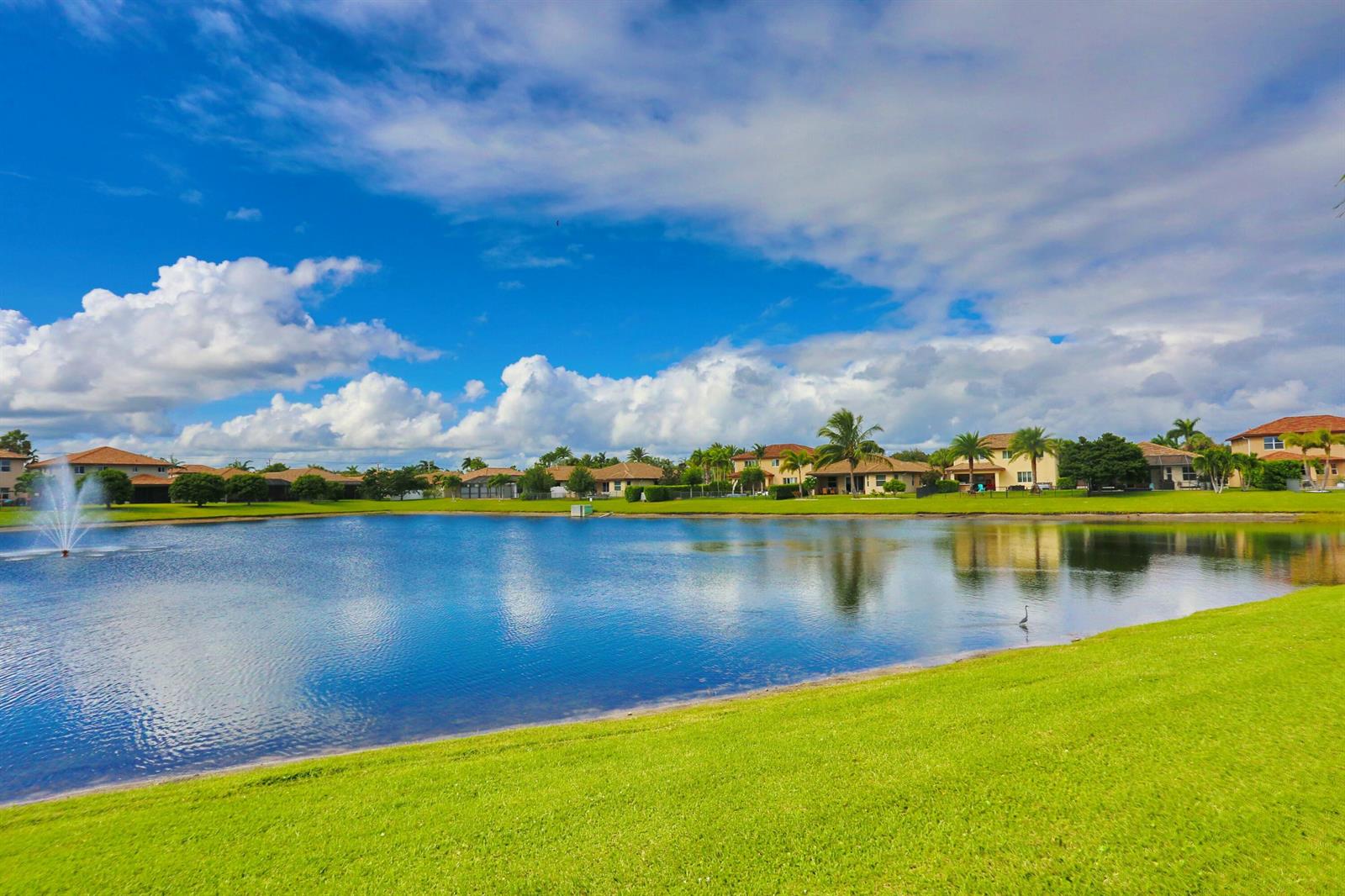 A lake with a fountain is surrounded by green grass and residential houses under clouds — 7171 Prudencia Drive, Lake Worth FL 33463, LAKE WORTH, FL