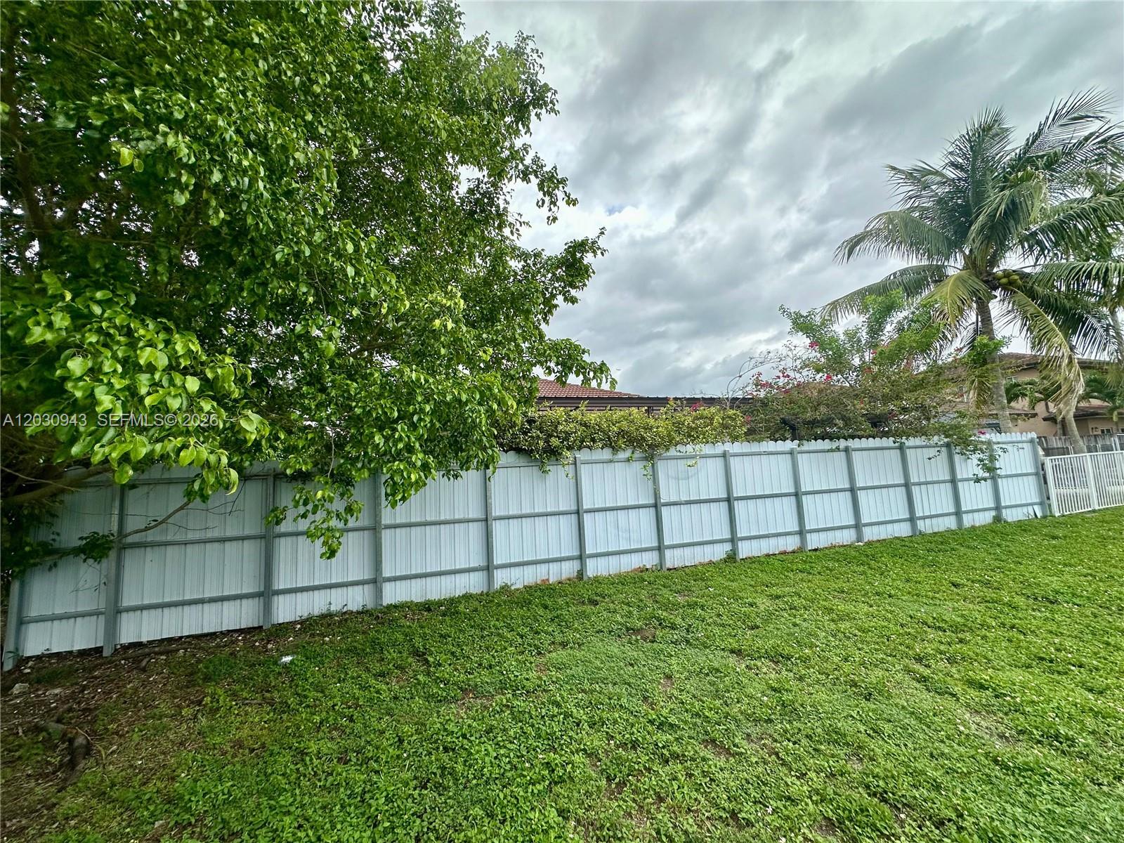 A white metal fence separates a grassy yard from trees and a cloudy sky — 15490 SW 10th St, Miami FL 33194, MIAMI, FL