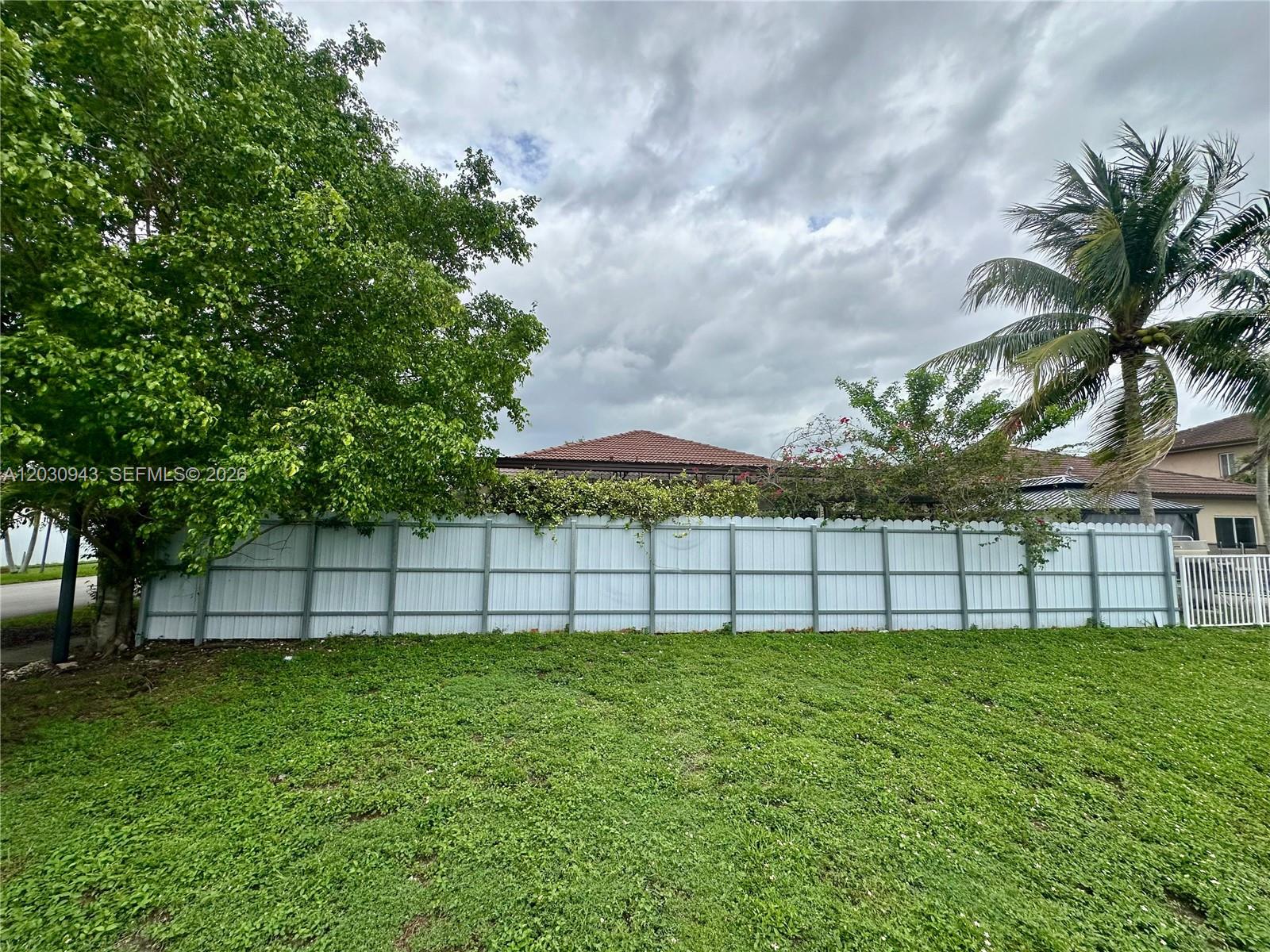 A white vinyl fence separates a grassy backyard from a house with a tiled roof — 15490 SW 10th St, Miami FL 33194, MIAMI, FL
