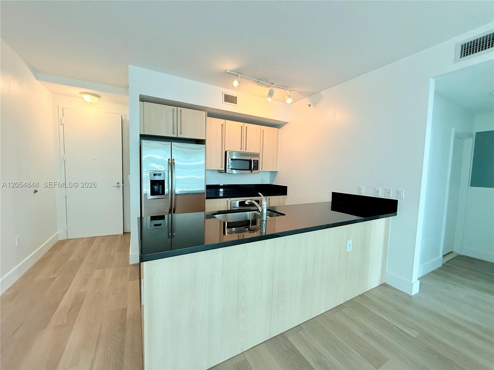 A kitchen features light wood cabinets, black countertops, stainless steel appliances, and wood flooring — MIAMI, FL