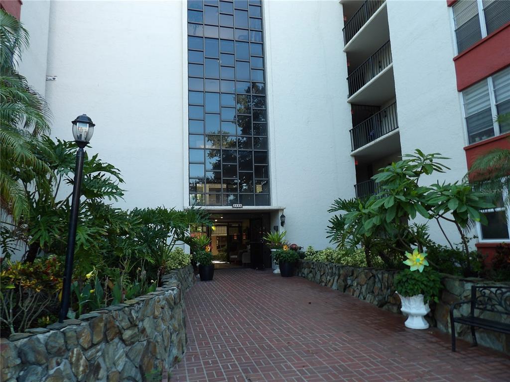 A brick walkway leads to the entrance of a multi-story apartment building with glass panels — 2699 SEVILLE BOULEVARD UNIT 802, CLEARWATER, FL