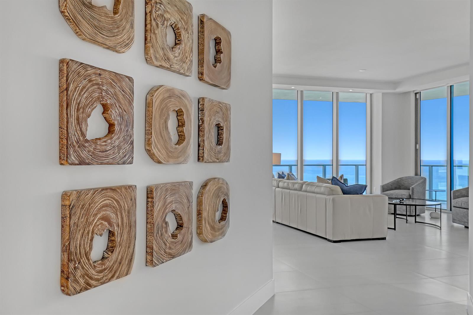 A hallway wall features nine wooden art pieces, leading into a bright living room overlooking ocean — 3100 N Ocean Drive # 1703, Riviera Beach FL 33404, RIVIERA BEACH, FL
