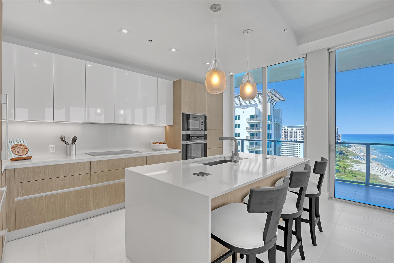 A modern kitchen features white glossy cabinets, light wood accents, a central island, and ocean views — 3100 N Ocean Drive # 1703, Riviera Beach FL 33404, RIVIERA BEACH, FL