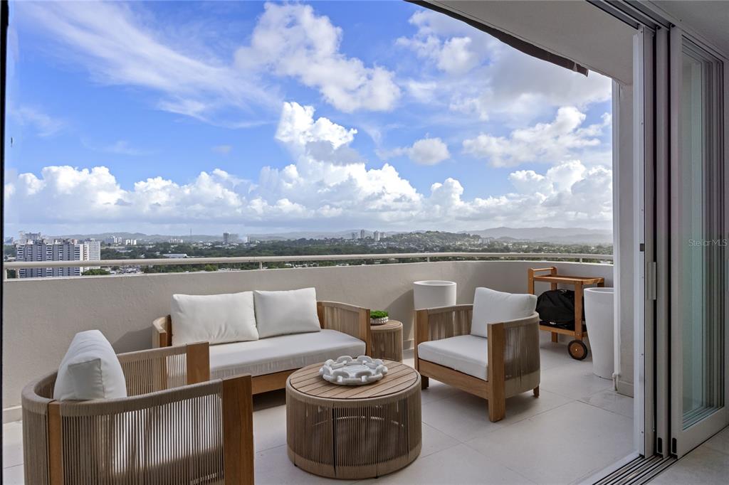 A balcony with modern outdoor furniture overlooks a city skyline under a cloudy blue sky — 225 ROAD NO 2, VILLA CAPARRA PLAZA UNIT PH-3, GUAYNABO, PR