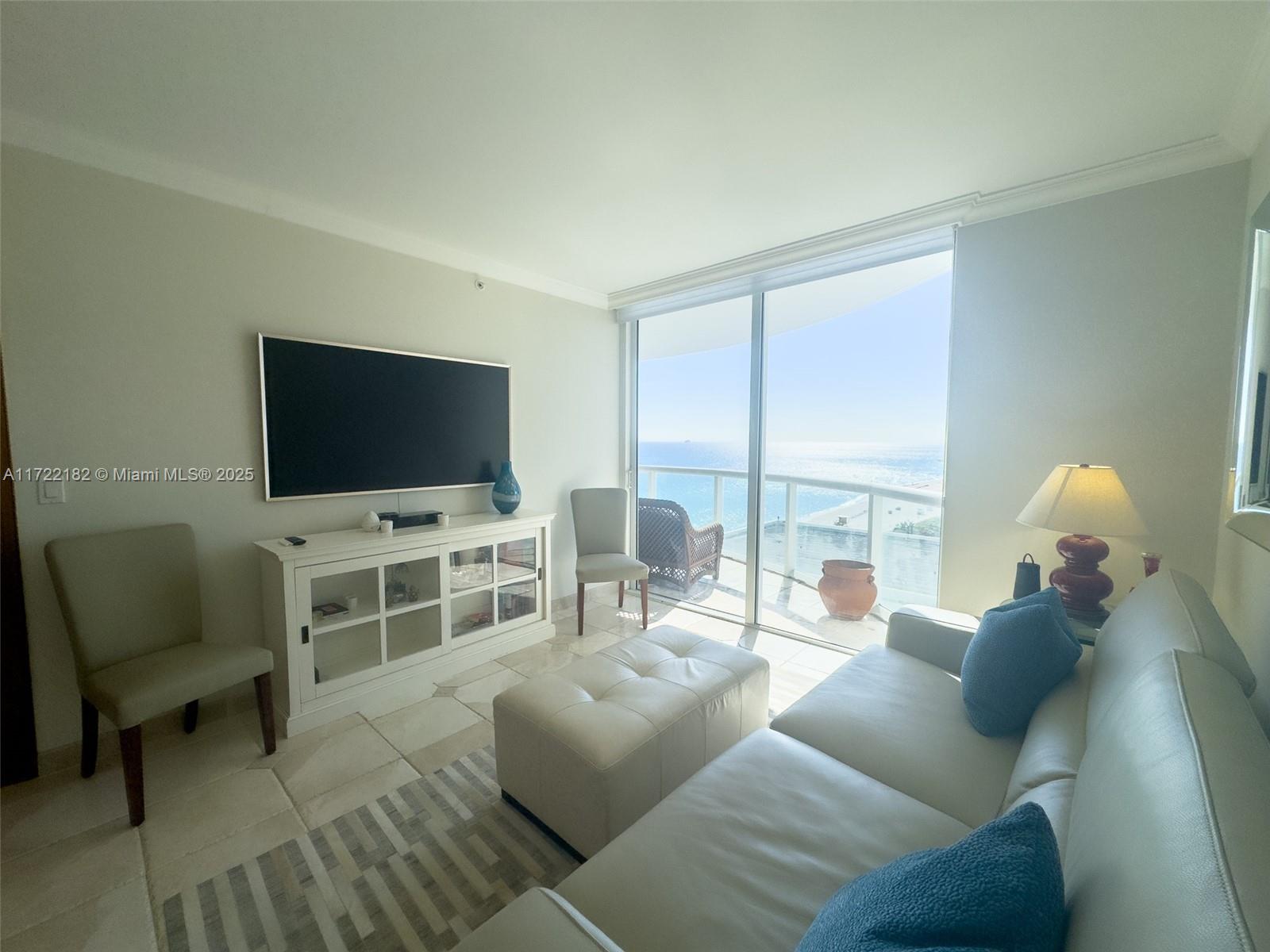 A living room features a white sofa, television, and floor-to-ceiling windows overlooking the ocean — 6365 Collins Ave # 1506, Miami Beach FL 33141, MIAMI BEACH, FL