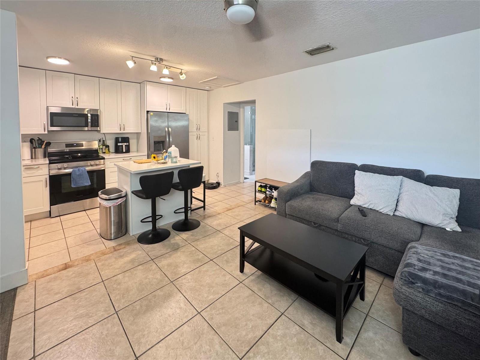 An open-concept kitchen and living area features white cabinetry, tile flooring, and a gray sofa — 151 SW 15th Street, Pompano Beach FL 33060, POMPANO BEACH, FL