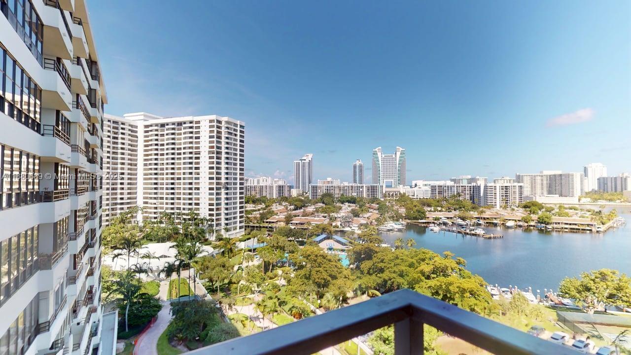 A high-angle view from a balcony overlooking a waterway, lush greenery, and distant city buildings — 500 Three Islands Blvd # 822, Hallandale Beach FL 33009, HALLANDALE BEACH, FL
