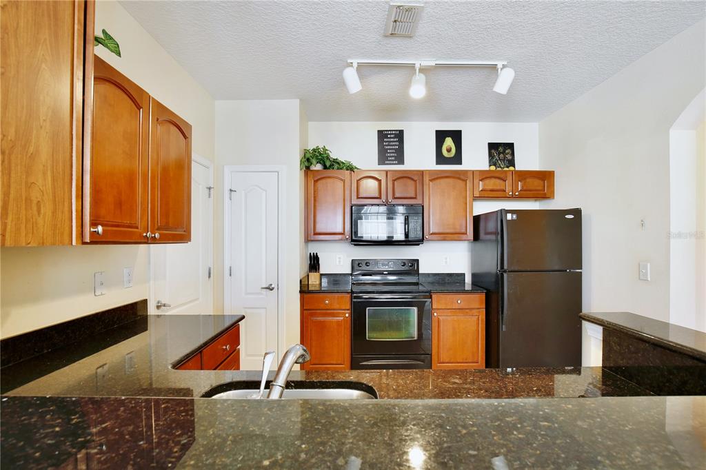 A kitchen features dark granite countertops, wooden cabinets, and black appliances under track lighting — 5255 CANE ISLAND LOOP UNIT 204, KISSIMMEE, FL