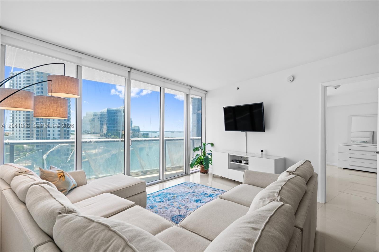 A modern living room features a beige sectional, a wall-mounted television, and floor-to-ceiling windows — 495 Brickell Ave, Miami FL 33131, MIAMI, FL