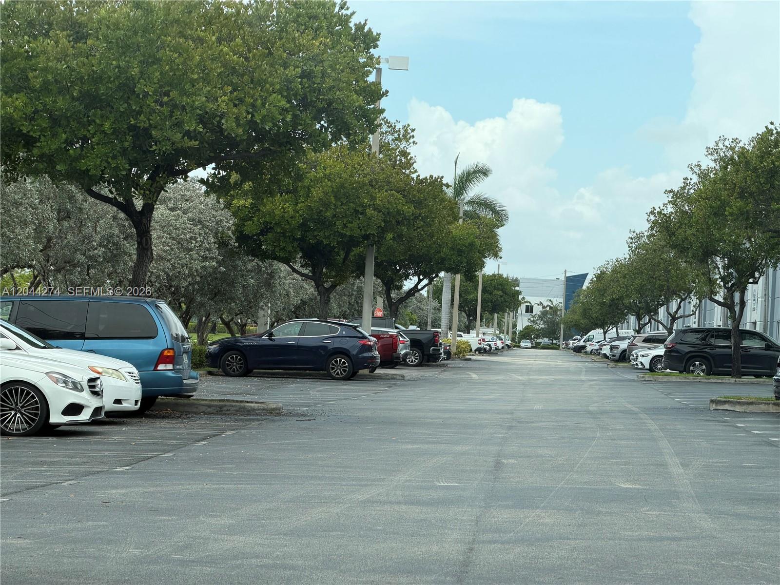 A paved parking lot is lined with parked vehicles and mature trees under a blue sky — 10871 NW 122nd St # 30, Medley FL 33178, MEDLEY, FL