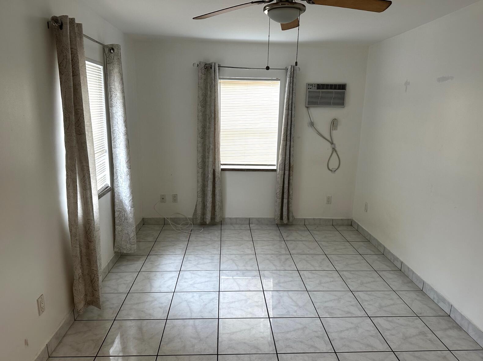 An empty room features white walls, light-colored tiled flooring, two windows with curtains, and an air conditioner — 31 SW 10th Street # 3, Dania Beach FL 33004, DANIA BEACH, FL