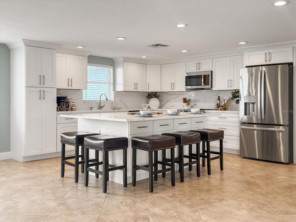 A white kitchen features a large island with four dark stools and stainless steel appliances — 5008 DORY DRIVE, NEW PORT RICHEY, FL