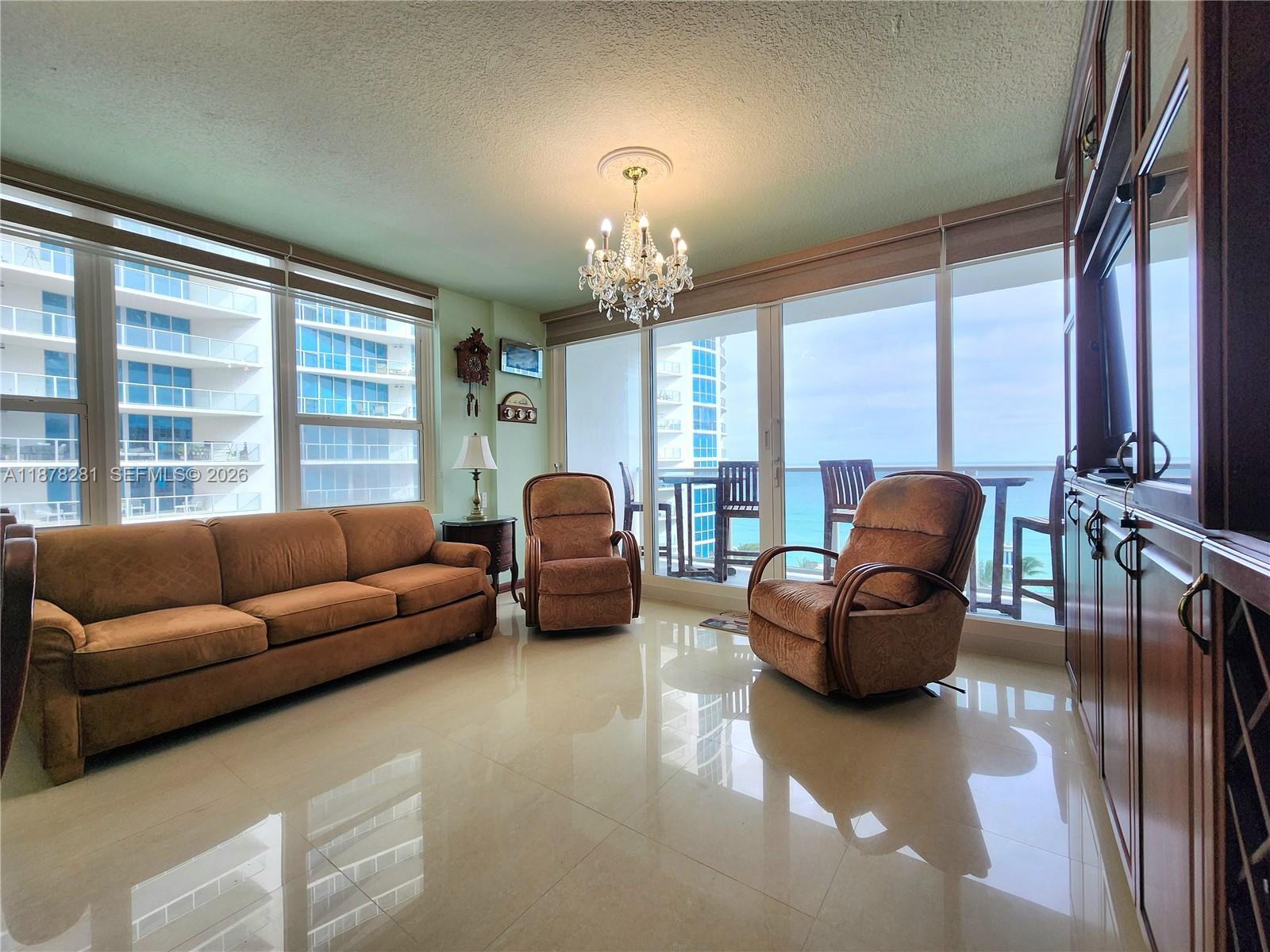 A living room features a brown sofa, two armchairs, a chandelier, and floor-to-ceiling ocean views — 3505 S Ocean Dr # 1006, Hollywood FL 33019, HOLLYWOOD, FL