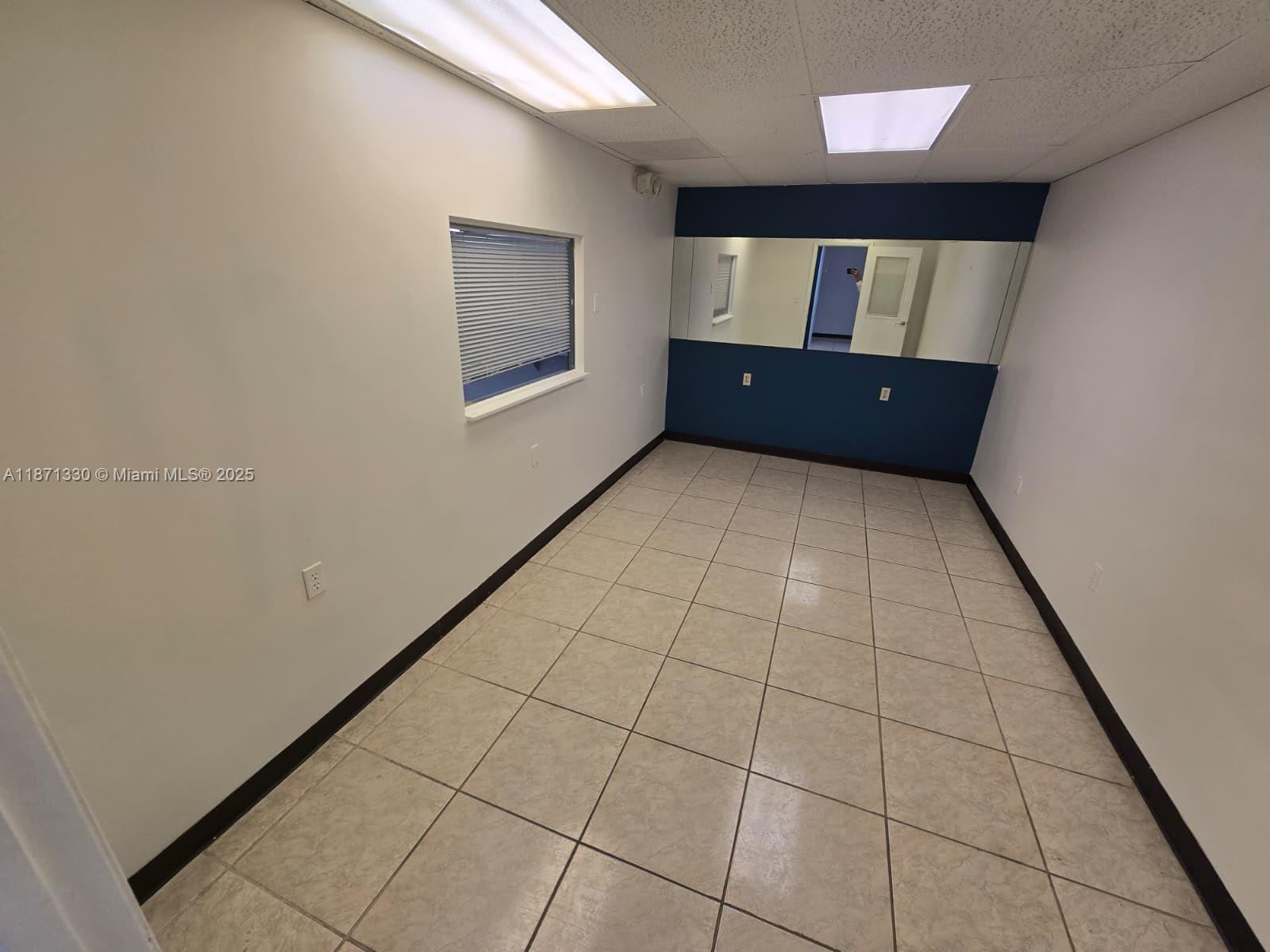 An empty room features light-colored tile flooring, white walls, a window, and a mirrored back wall — 2501 N State Road 7 # 216, Margate FL 33063, MARGATE, FL