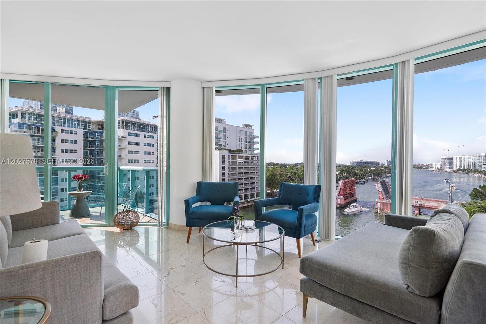 A living room with gray sofas, blue armchairs, and floor-to-ceiling windows overlooking a waterway — 6000 Indian Creek Dr # 8B, Miami Beach FL 33140, MIAMI BEACH, FL