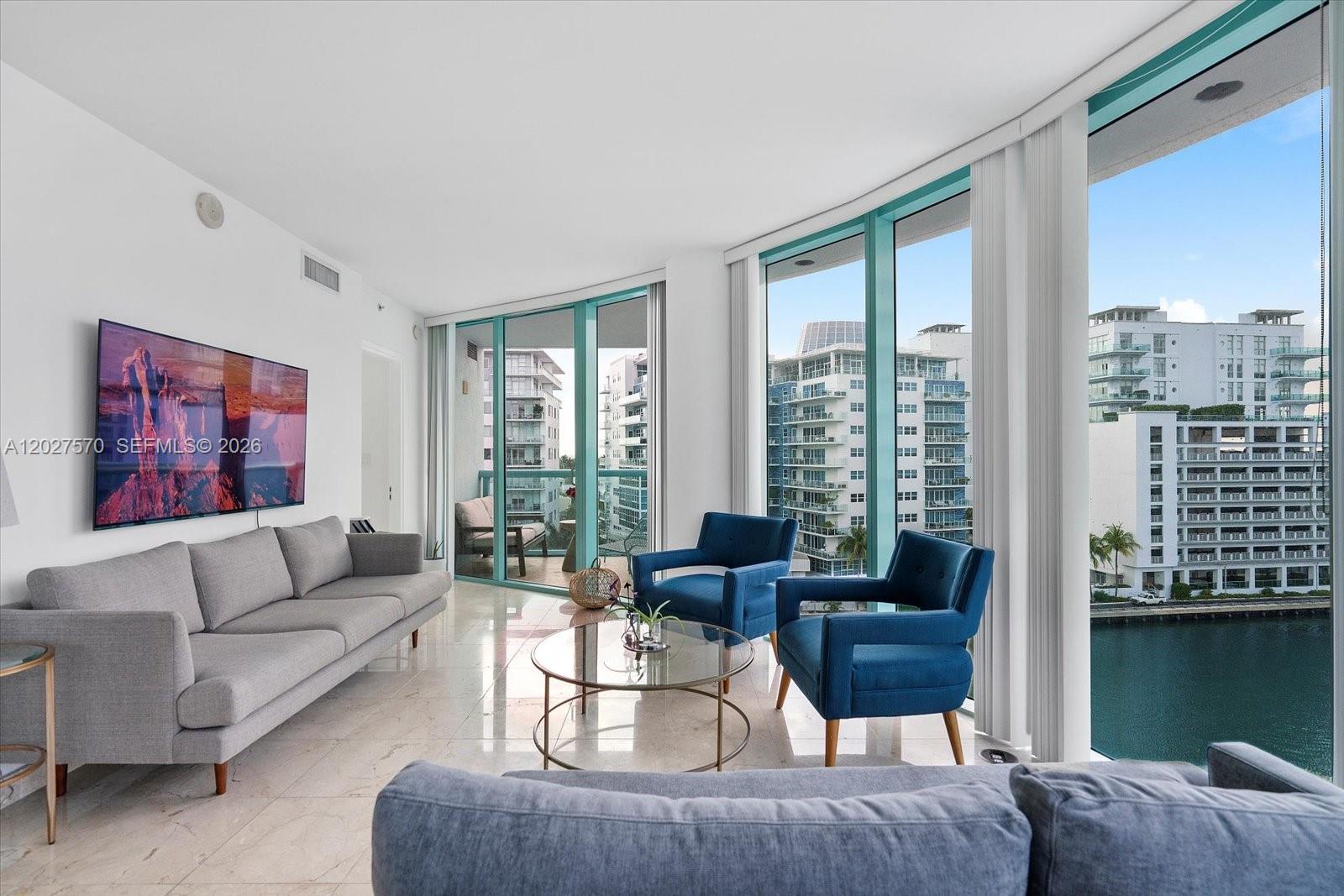 A living room features a gray sofa, two blue armchairs, and floor-to-ceiling windows overlooking water — 6000 Indian Creek Dr # 8B, Miami Beach FL 33140, MIAMI BEACH, FL