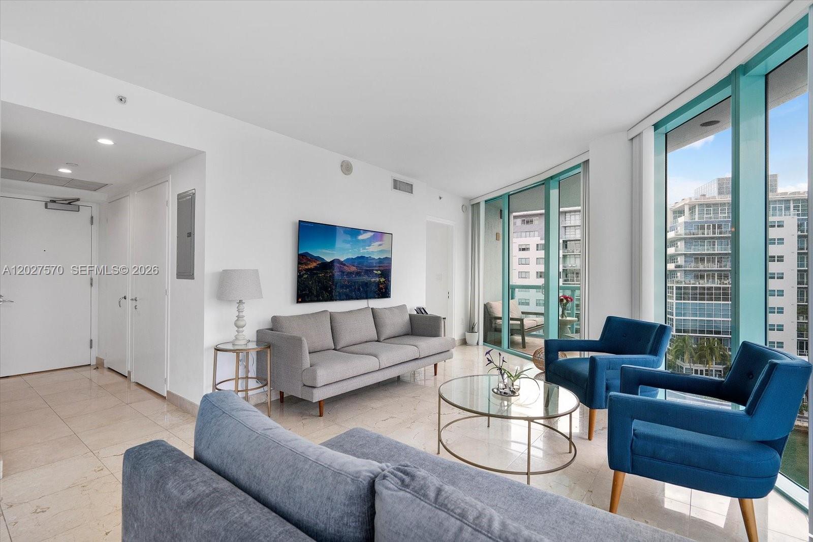 A living room features a grey sofa, two blue armchairs, and floor-to-ceiling windows — 6000 Indian Creek Dr # 8B, Miami Beach FL 33140, MIAMI BEACH, FL