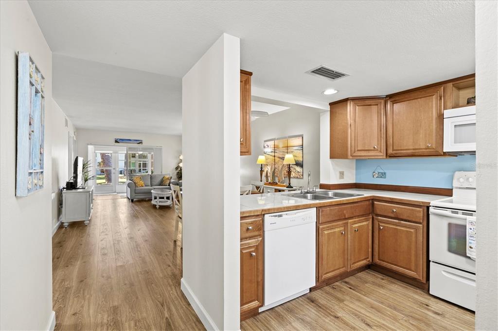 A kitchen with wood cabinets and white appliances opens into a living area with flooring — 404 PINEBROOK CRESCENT UNIT 4, VENICE, FL