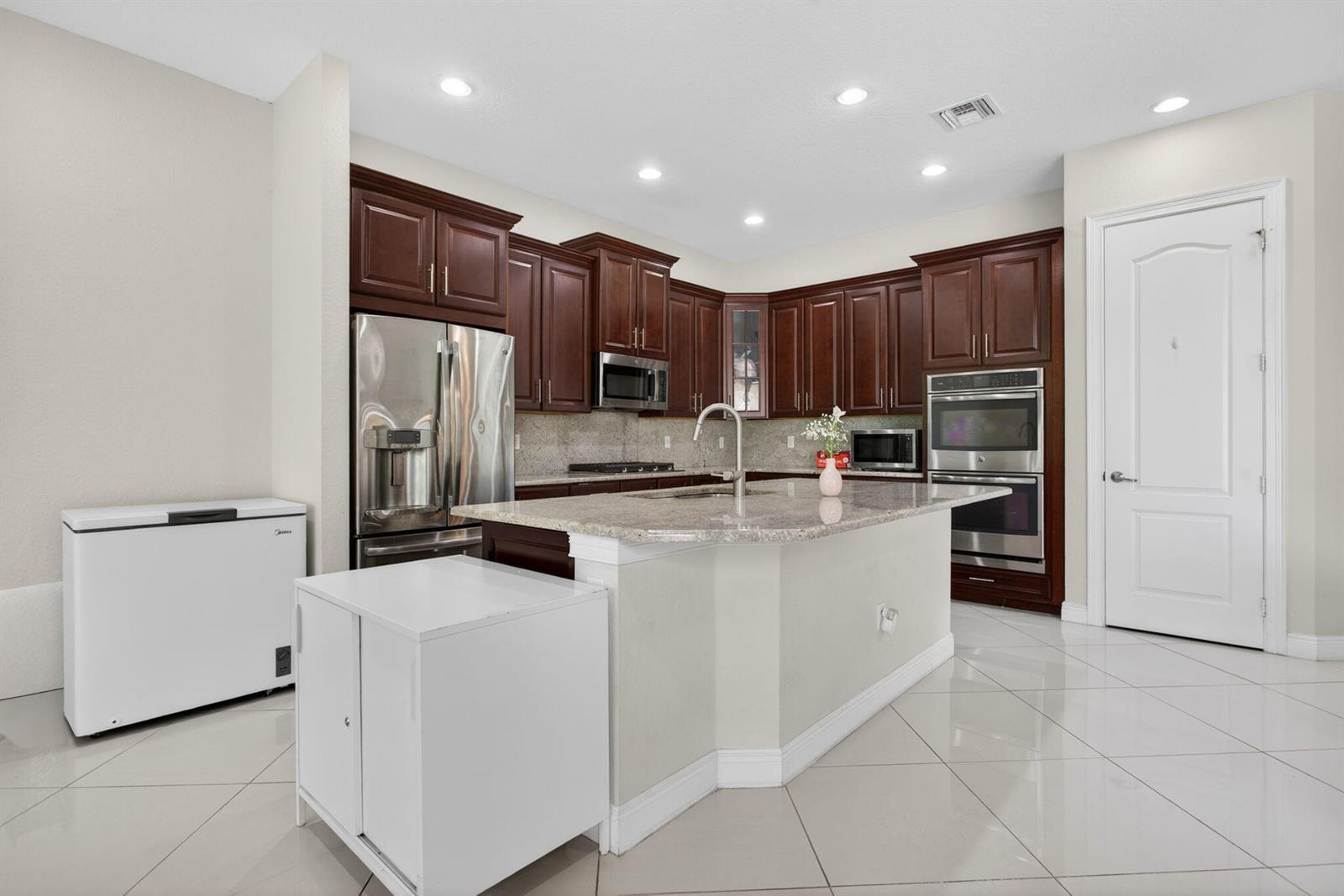 A kitchen features dark wood cabinets, granite countertops, stainless steel appliances, and white tile flooring — 8850 Miralago Way, Parkland FL 33076, PARKLAND, FL