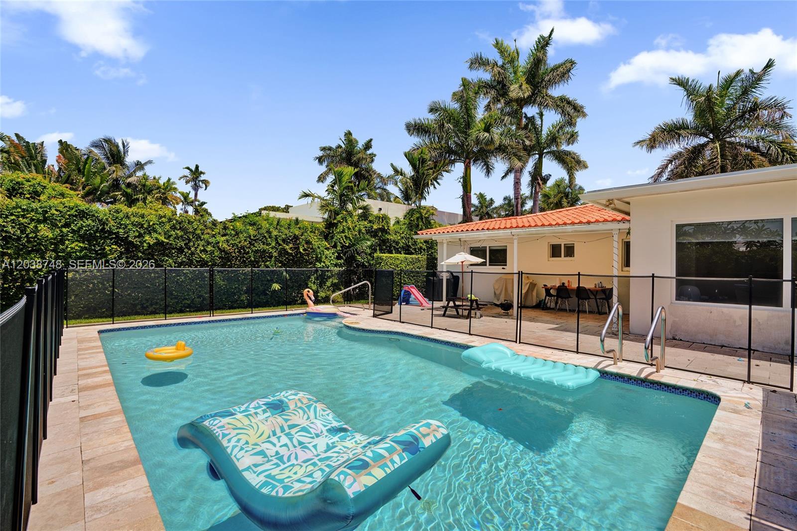 A backyard swimming pool with floating rafts is enclosed by a black fence and palm trees — 2025 Alamanda Dr, North Miami FL 33181, NORTH MIAMI, FL