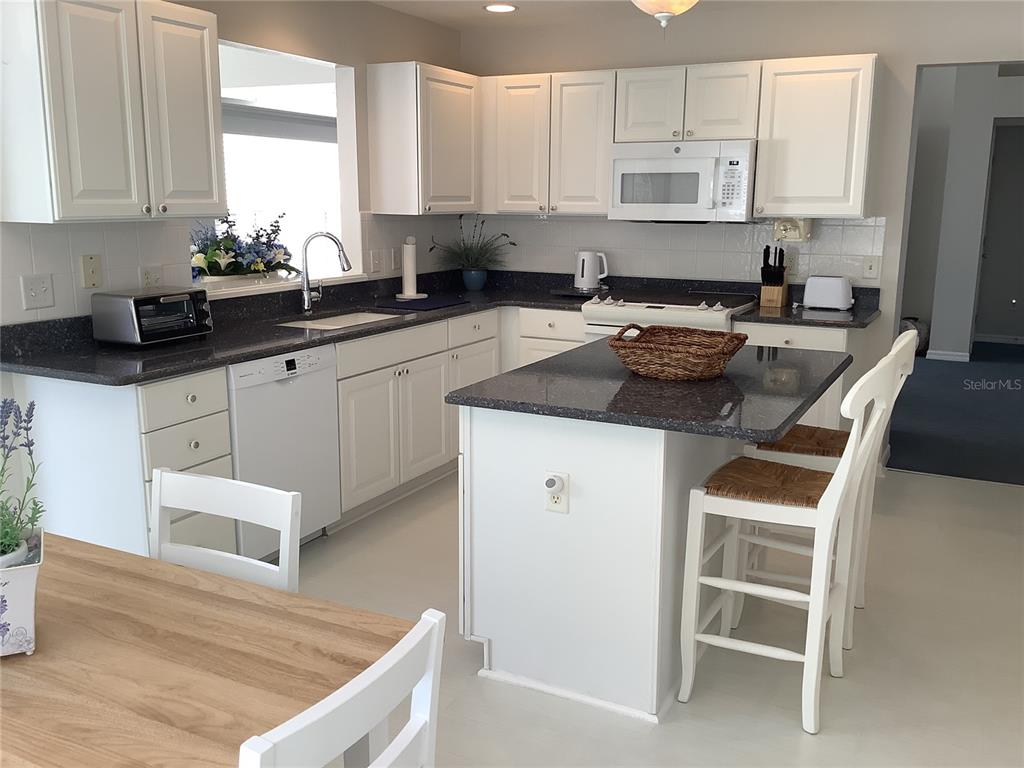 A kitchen features white cabinets, dark granite countertops, a center island, and white appliances — 890 HAYNESVILLE WAY, THE VILLAGES, FL