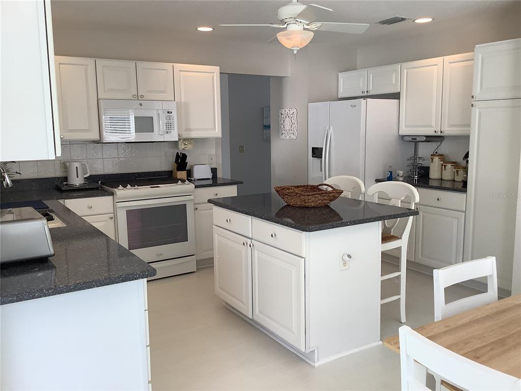 A kitchen features white cabinetry, dark granite countertops, a central island, and white appliances — 890 HAYNESVILLE WAY, THE VILLAGES, FL